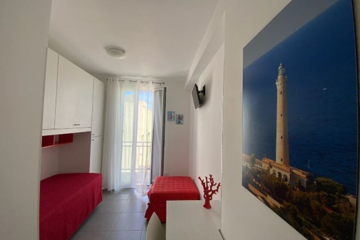 Una stanza modesta con un letto a castello, un tavolino, una sedia, un quadro raffigurante un faro e una porta che dà su un'altra stanza.