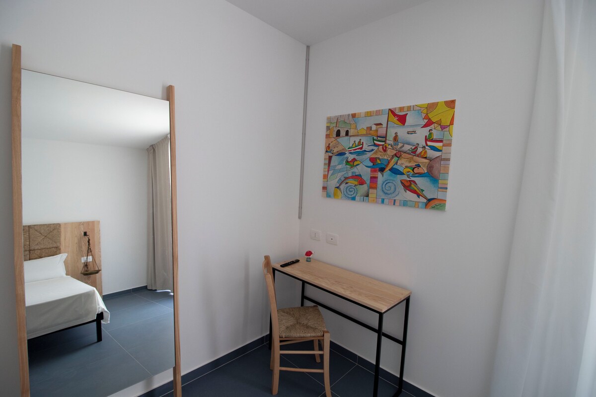 A bedroom with a bed, a desk, a chair, a mirror, and a colorful painting on the wall.