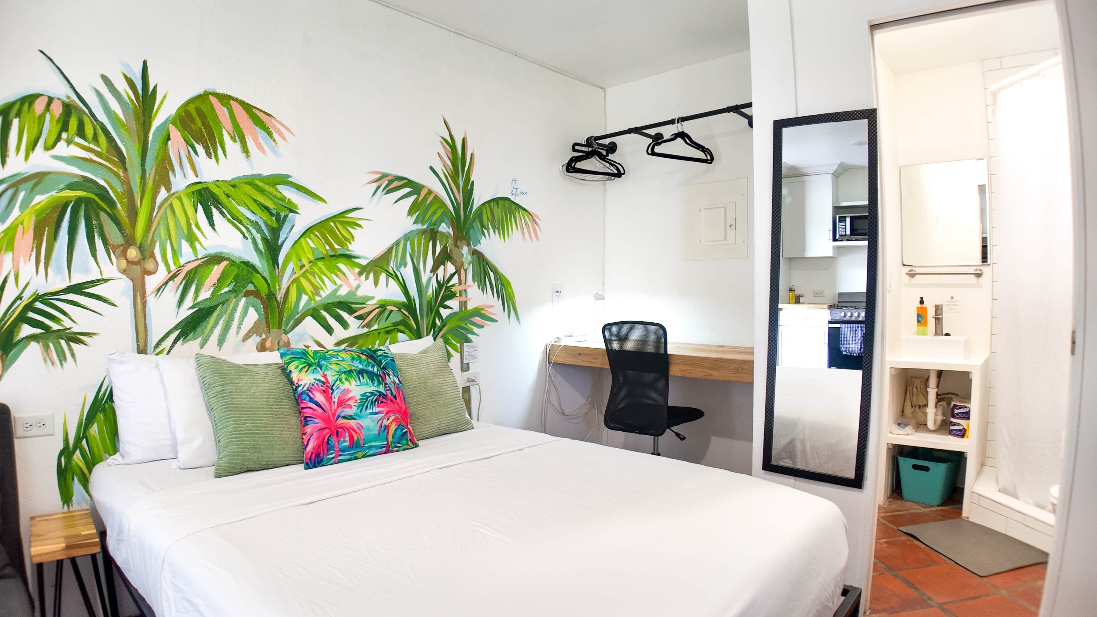 A hotel room with a bed, a desk, a chair, a mirror, and a kitchen area visible through an open door.