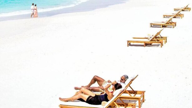 Una spiaggia con sabbia bianca e acqua cristallina. Sono presenti sedie a sdraio in legno e una coppia che passeggia sulla spiaggia.