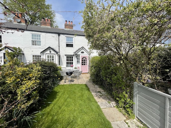 Une maison blanche à deux étages, avec une porte rose et un petit jardin comprenant une pelouse et un espace barbecue.