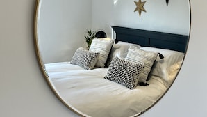 Een slaapkamer met een bed, kussens, een nachtkastje en een spiegel aan de muur.