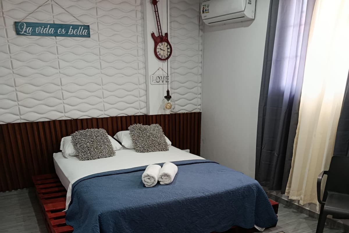 A neatly made bed with a blue bedspread and two pillows, a wall clock, and a guitar hanging on the wall.