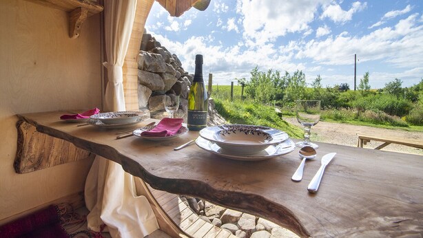 Mesa de madera preparada para comer, con platos, cubiertos y una botella de vino, con vistas a un paisaje natural.