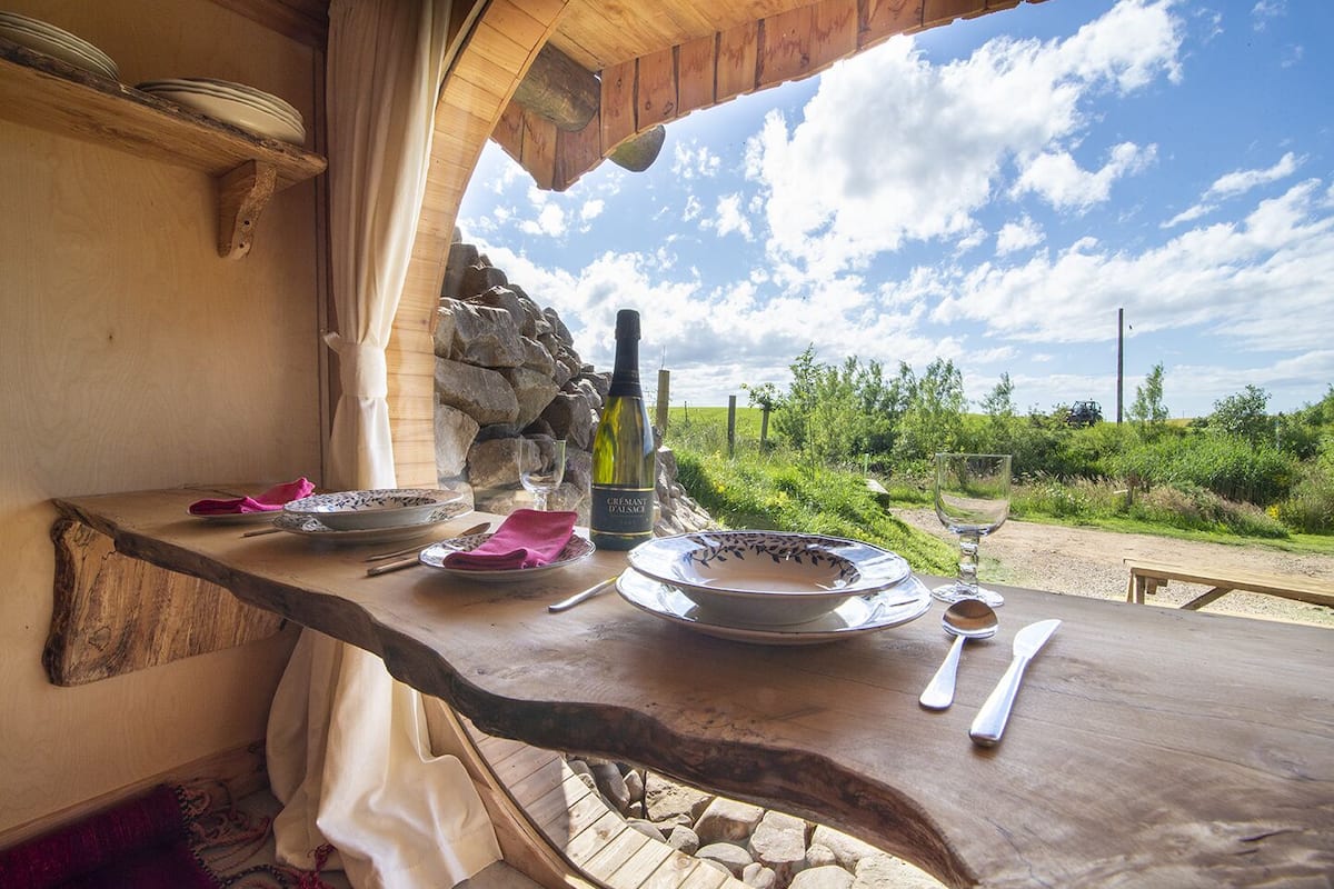A wooden table set for dining with plates, cutlery, and a bottle of wine, overlooking a natural landscape.