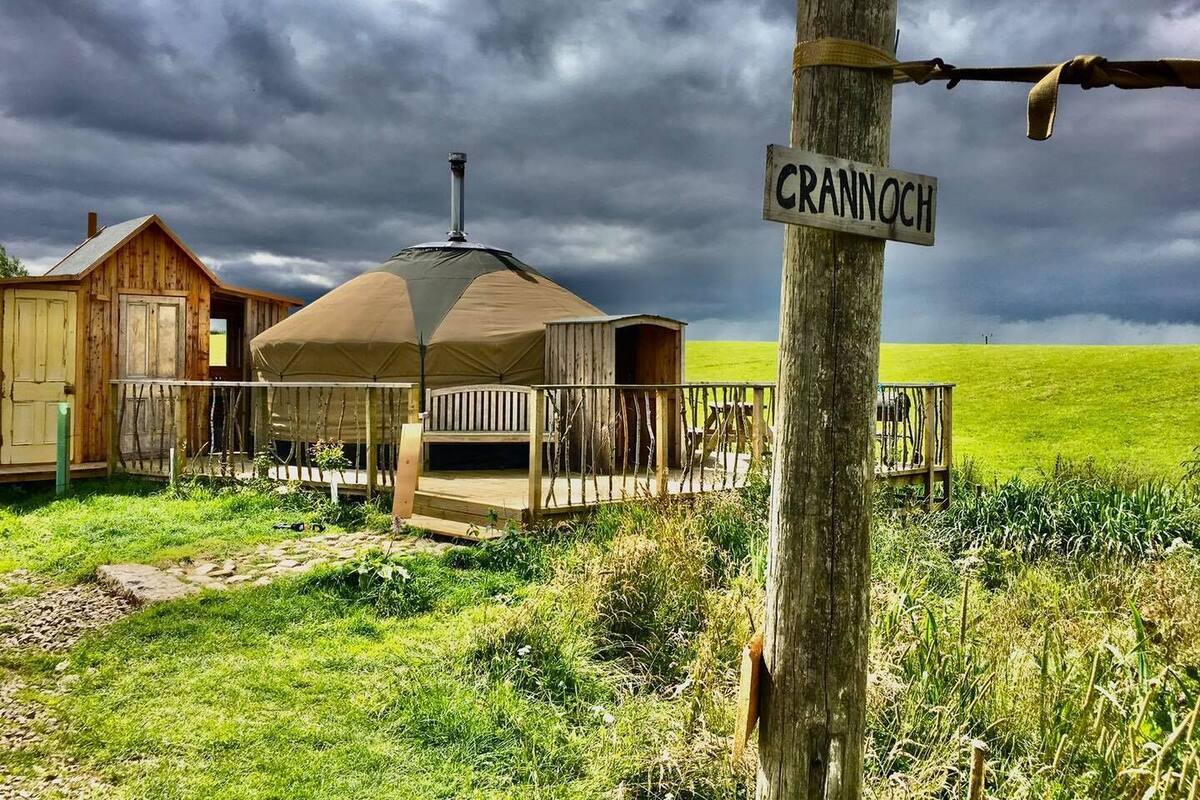 A rustic wooden structure with a covered seating area, a sign reading "CRANNOCCH," and a grassy field in the background.