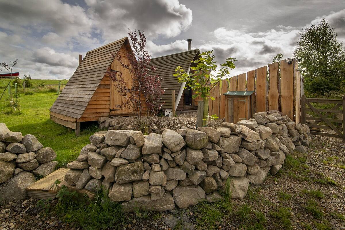 A rustic wooden structure with a sloped roof, surrounded by a stone wall and a wooden fence.