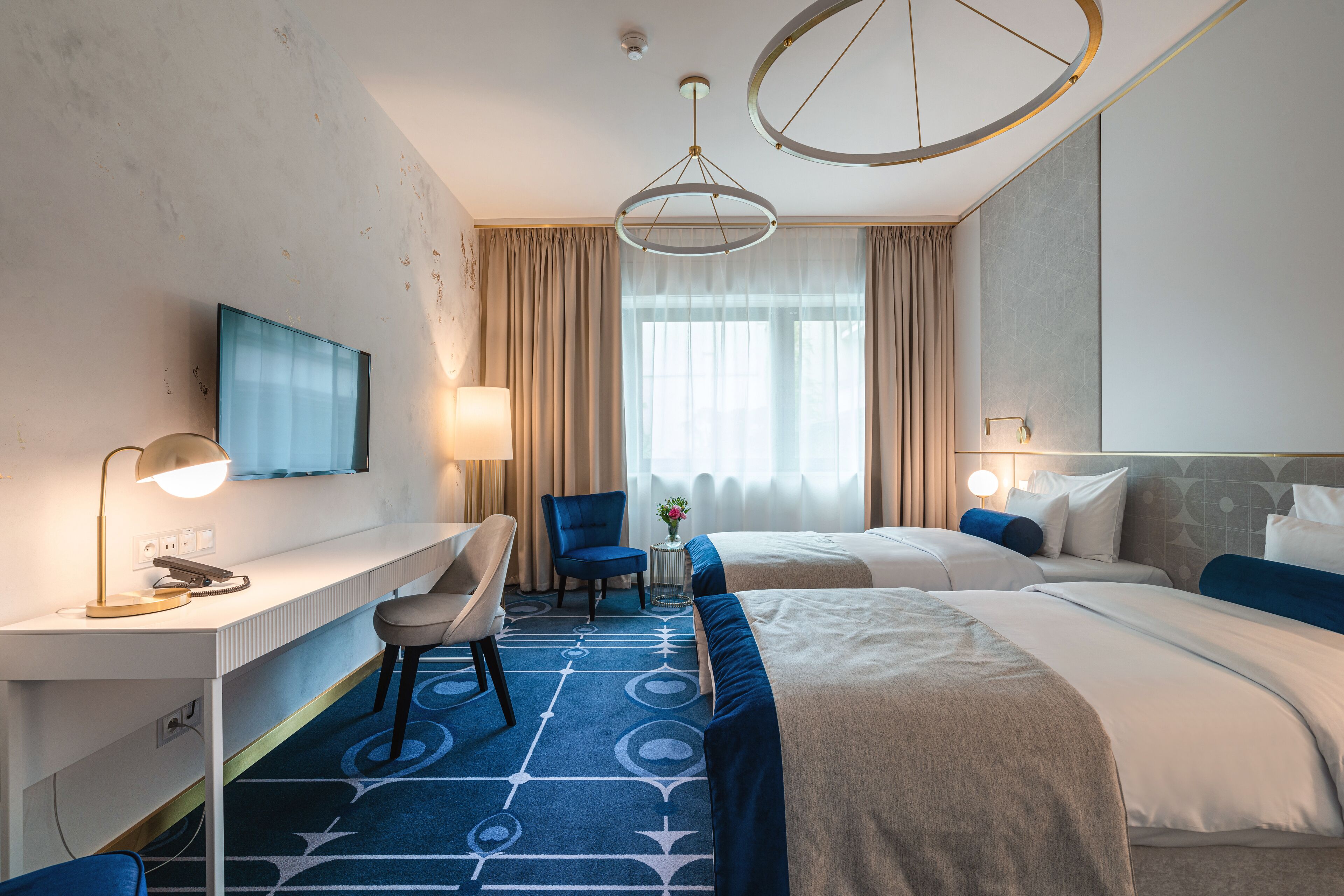 Une chambre d’hôtel avec deux lits, un bureau, une chaise, une télévision et une grande fenêtre avec des rideaux.