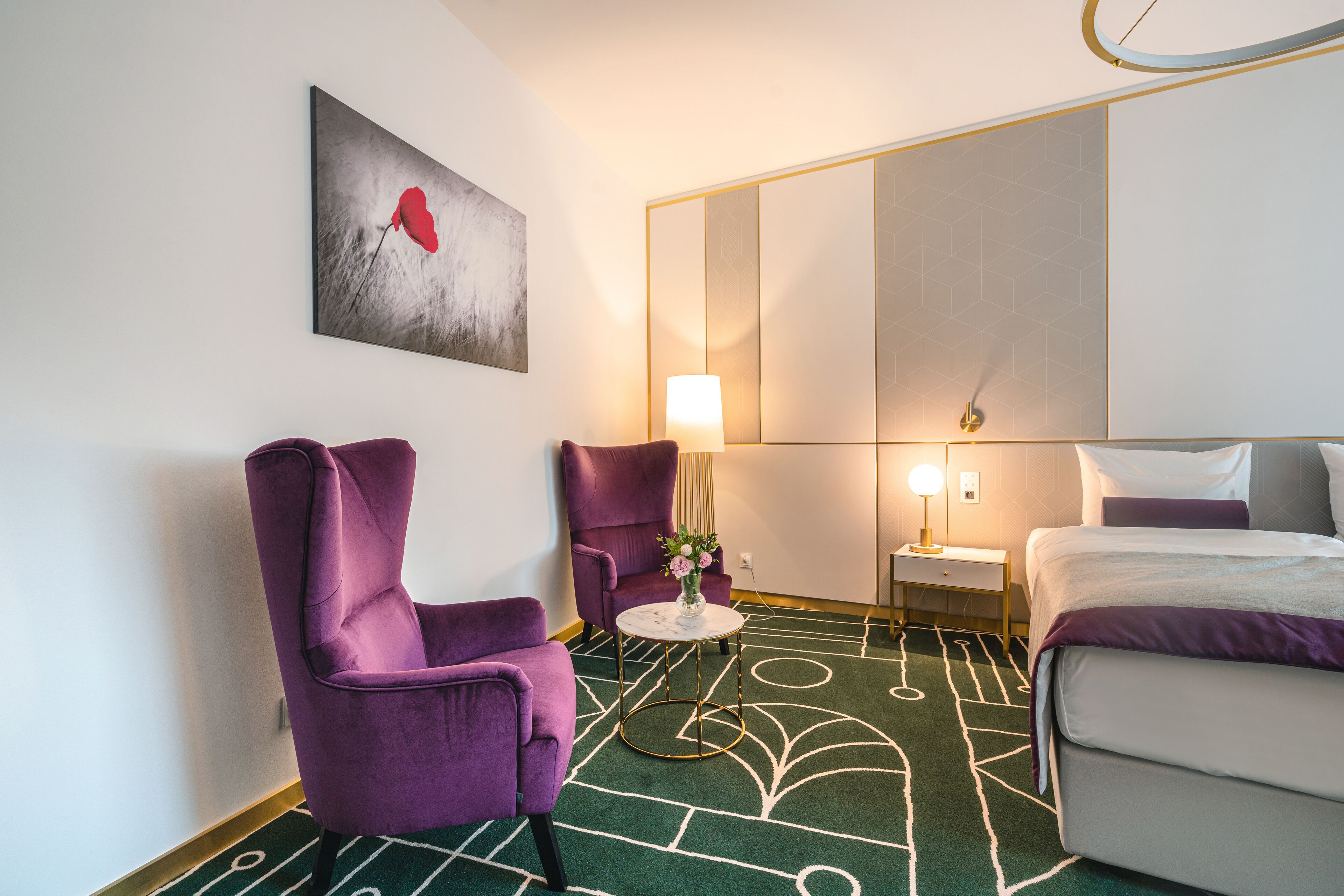Une chambre d’hôtel avec un lit, deux fauteuils violets, une petite table ronde ornée d’un vase de fleurs, une table de chevet avec une lampe et un grand miroir.