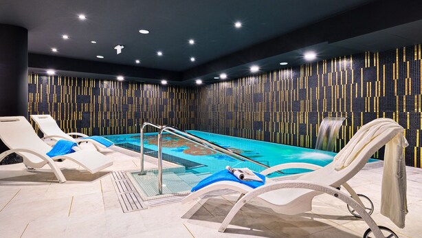 Een modern binnenzwembad met loungebedden, een zwembad met waterval en een betegelde muur.