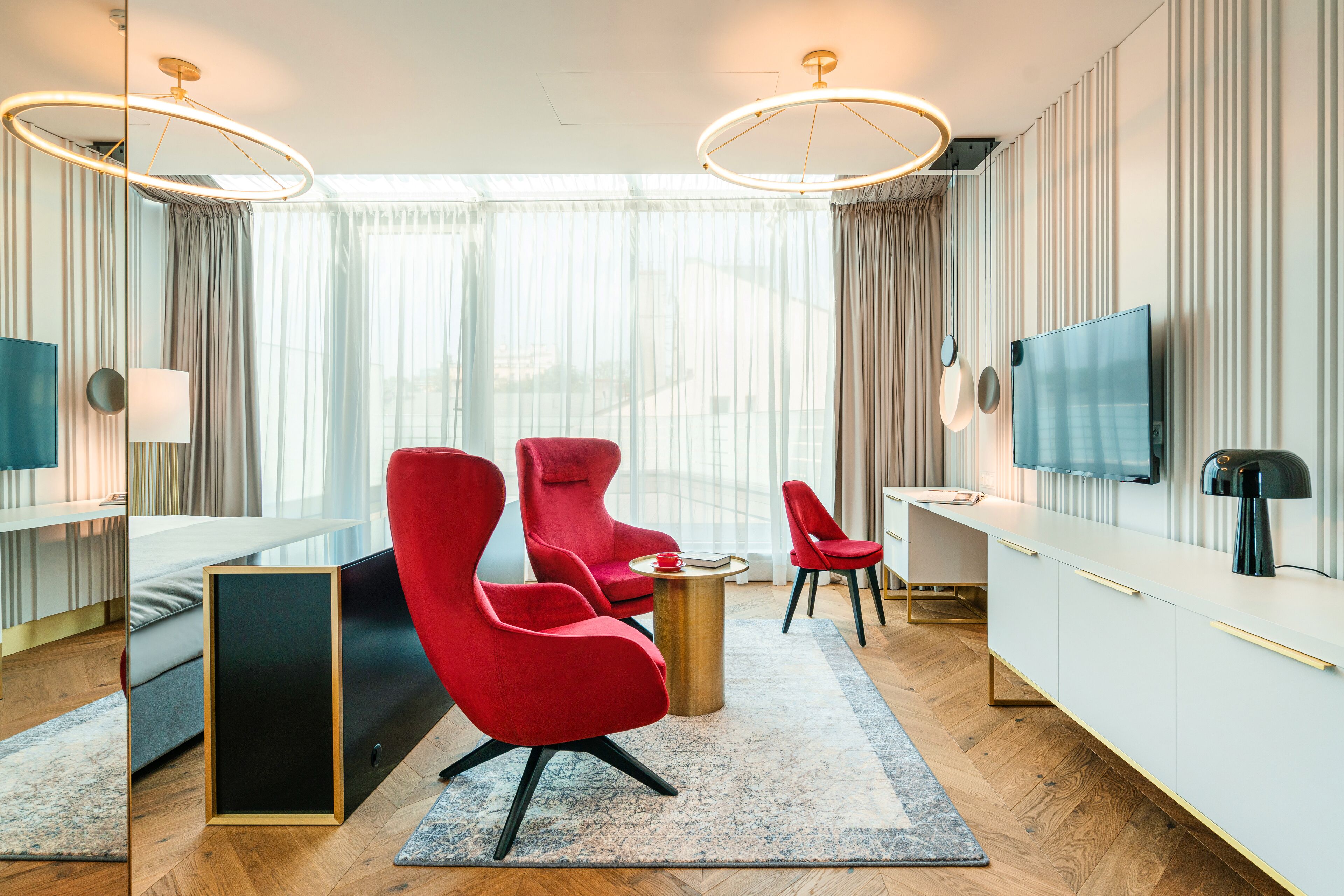 A modern hotel room with a large bed, a desk, two red chairs, a TV, and a large window with curtains.