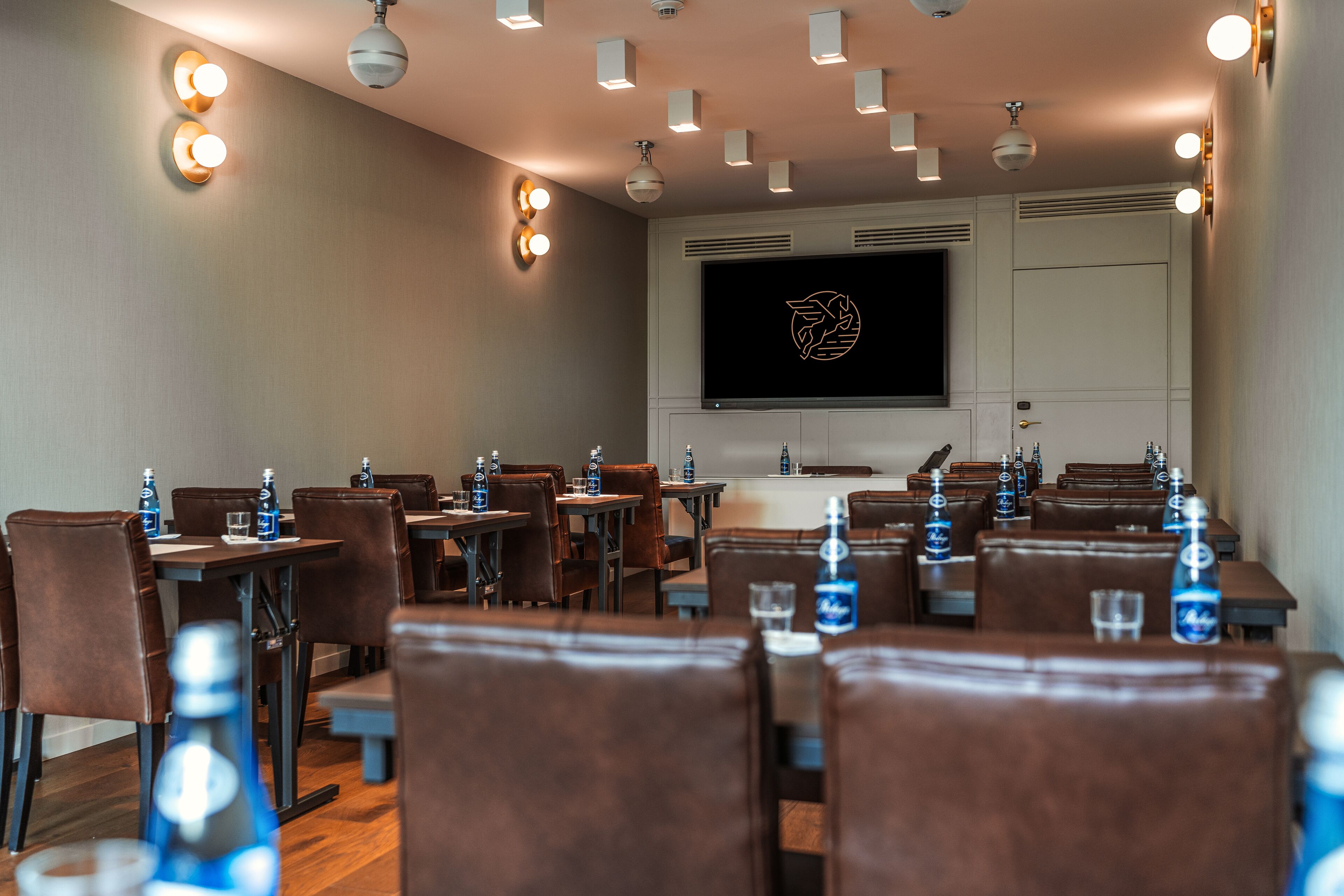 Ein Konferenzraum mit Tischen, Stühlen, einem großen Bildschirm und Deckenleuchten.