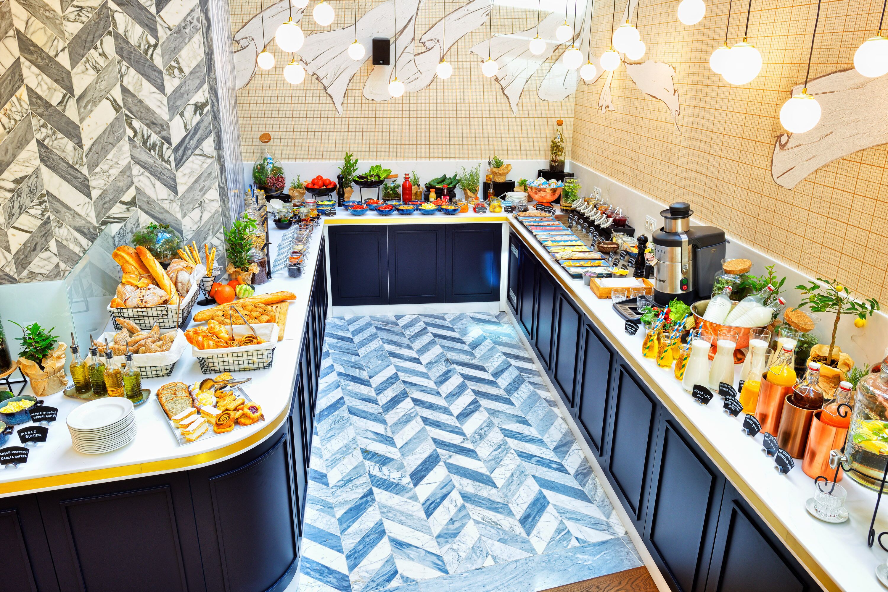 A buffet setup with various food items and beverages, a patterned floor, and a modern kitchen area with hanging lights.