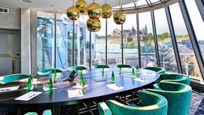 Een congreszaal met panoramisch stadszicht, een ronde tafel, teal stoelen en gouden, bolvormige lampen.