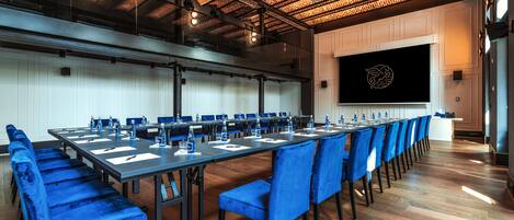 A conference room with a long table, blue chairs, and a projector screen.