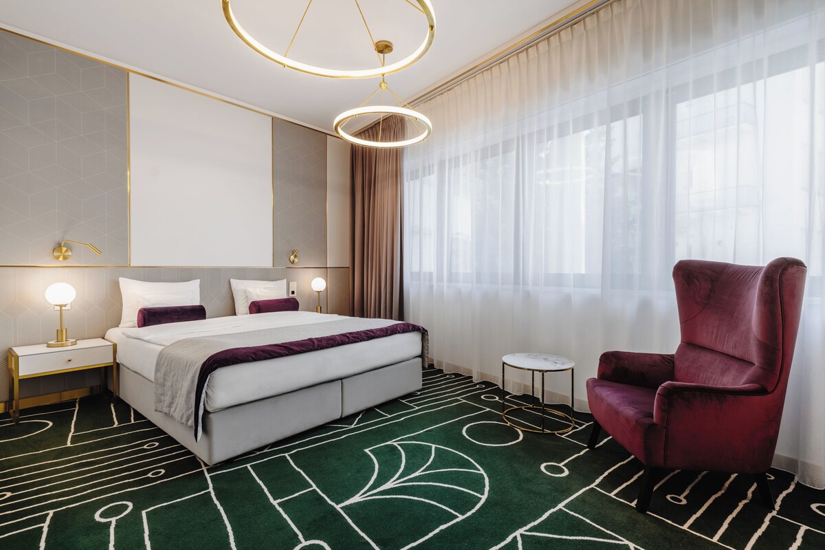 A hotel room with a bed, a red armchair, a small table, and a large window with sheer curtains.
