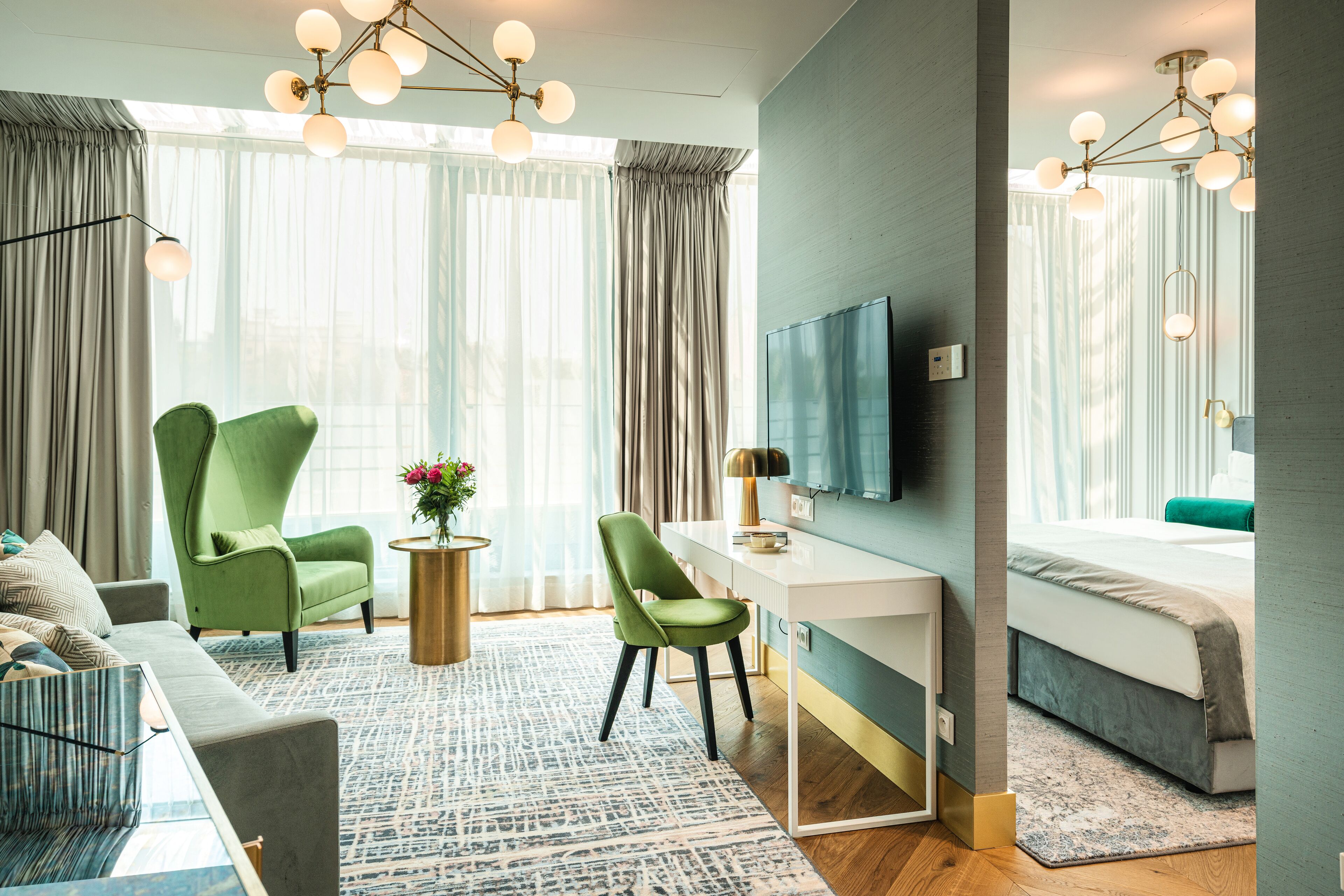 A modern hotel room with a green armchair, a desk, a flat-screen TV, and a large window with sheer curtains.