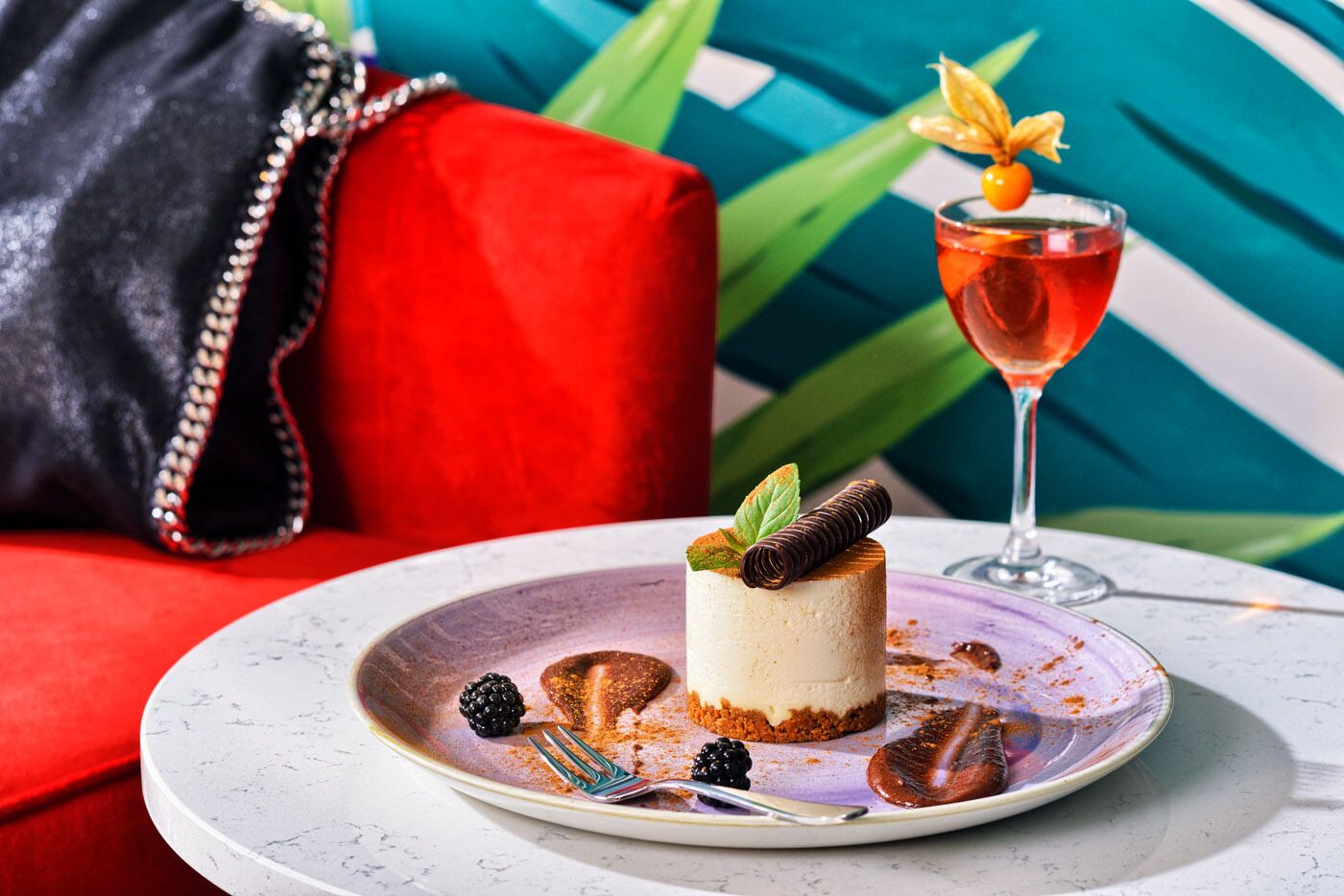 A dessert on a plate with a fork, a glass of red drink, and a black handbag on a red couch.