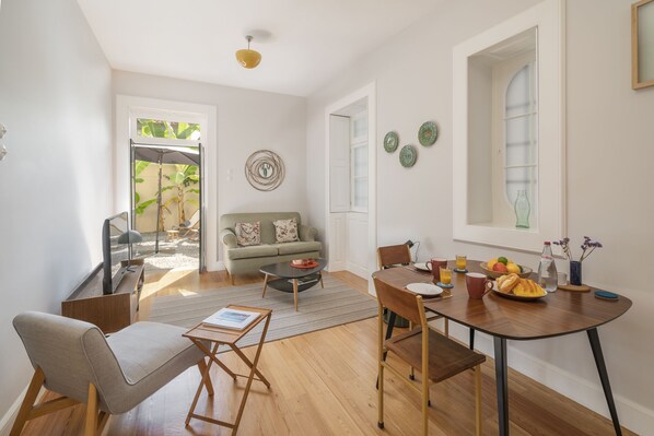 A modern living room with a sofa, coffee table, dining table, and chairs. There is a television, a small table with a bowl of fruit, and a vase with flowers.