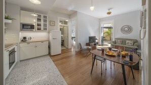 A modern kitchen with white cabinets, a microwave, and a refrigerator. There is a dining area with a table set for two, a sofa, and a TV. - Chill Garden Apartment (Funchal)