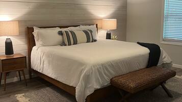 A bedroom with a wooden headboard, a large bed with white bedding, a bedside table with a lamp, a bench, a ceiling fan, and a window with blinds.