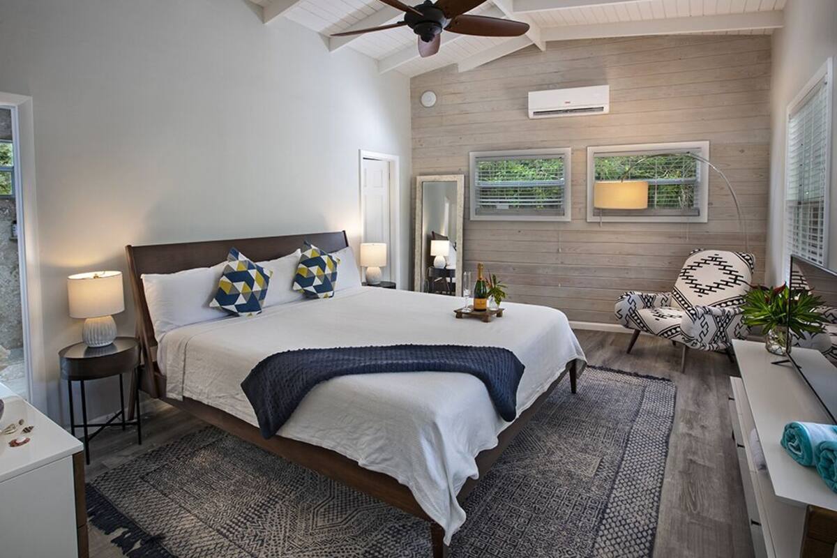 A bedroom with a large bed, a ceiling fan, a bedside table with a lamp, a chair, and a window with blinds.