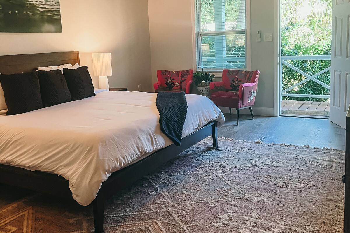 A bedroom with a large bed, a ceiling fan, a window with blinds, and a view of greenery outside.