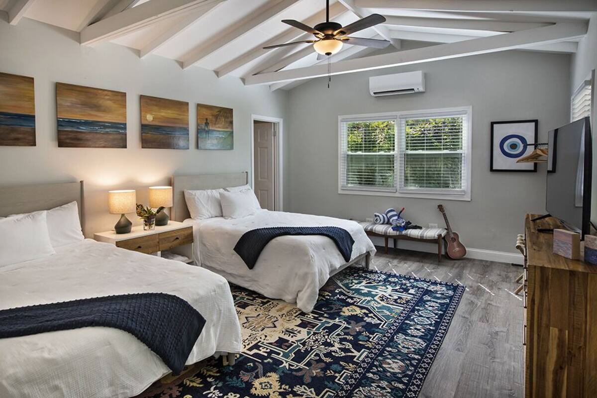 A bedroom with two beds, a ceiling fan, artwork on the walls, a window with blinds, and a guitar on a bench.