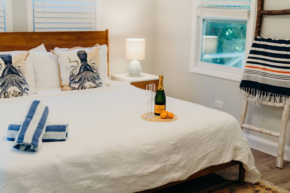 A neatly made bed with a white bedspread, two pillows with octopus designs, a bottle of champagne, two glasses, and two oranges on a wooden tray.