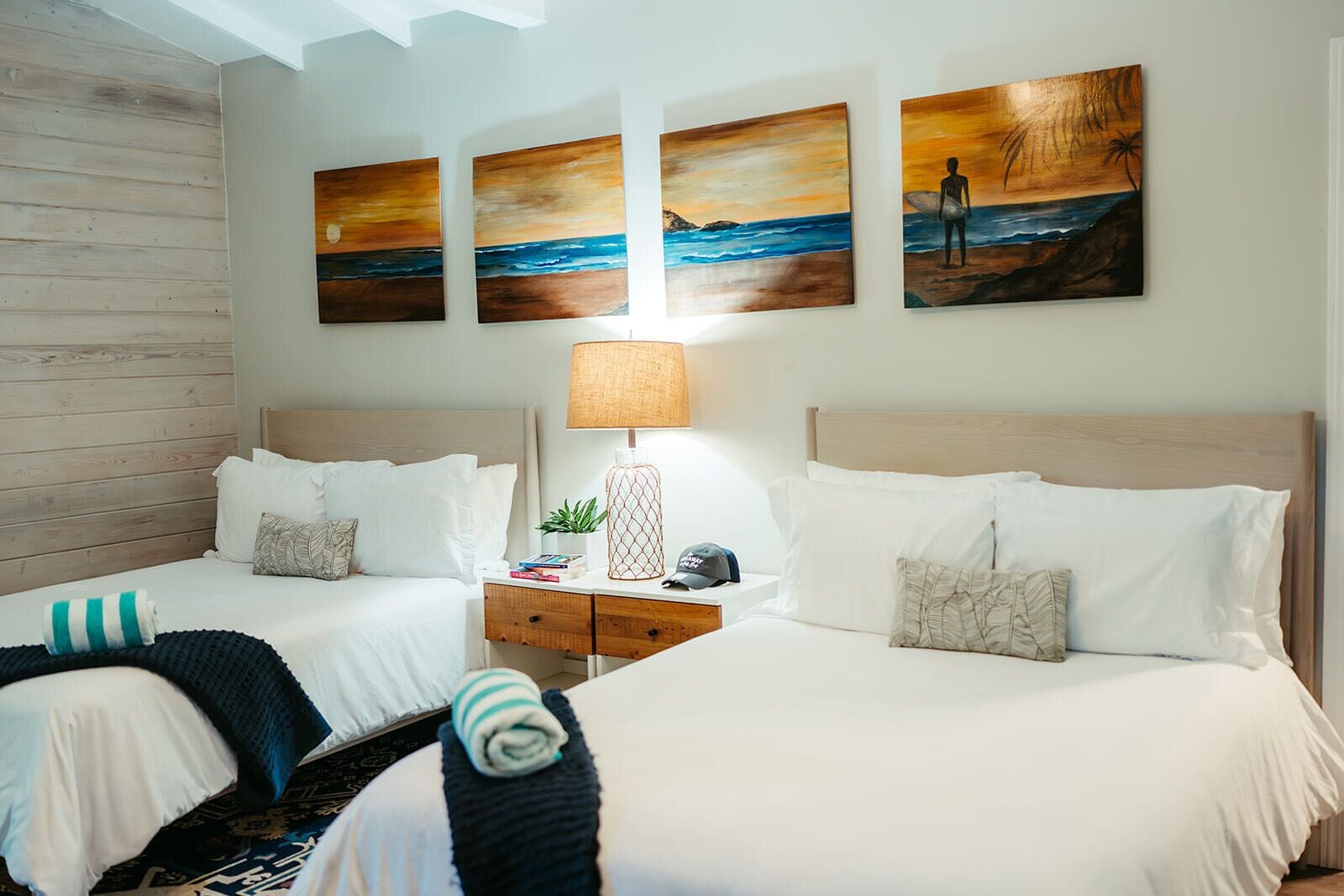 A hotel room with two beds, a nightstand with a lamp, and three paintings on the wall.