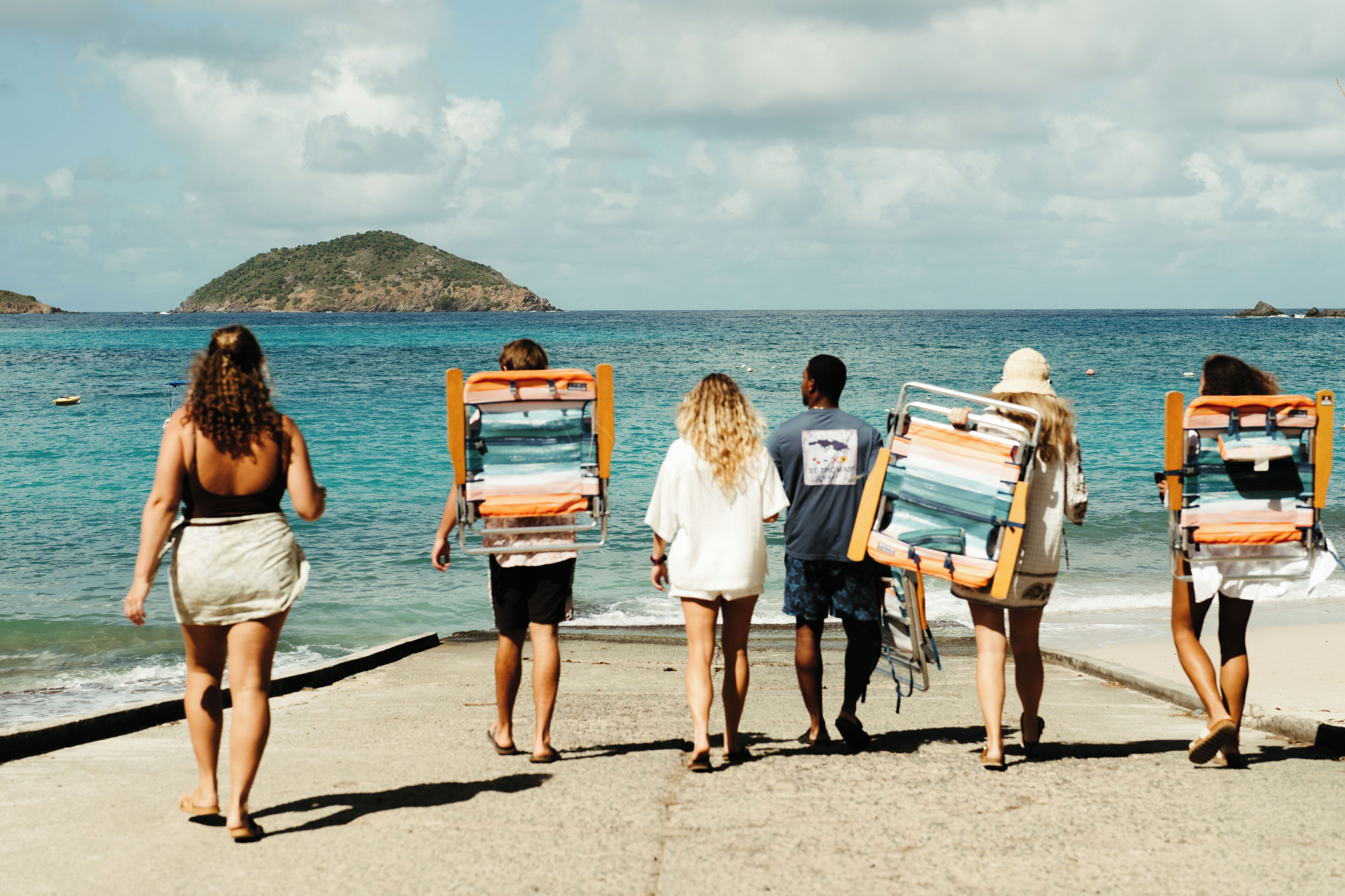 A group of people walking along a beach with beach chairs.