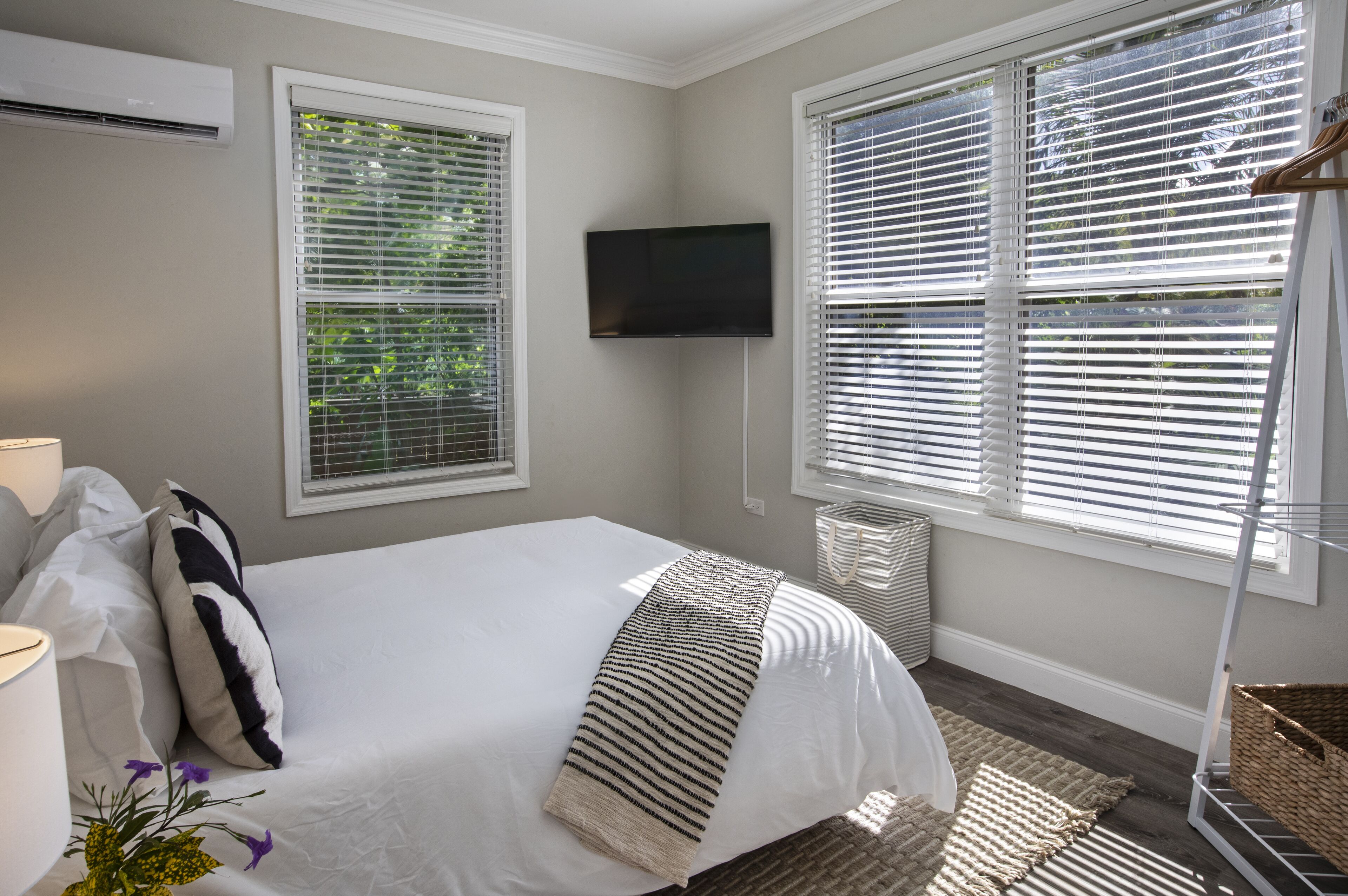 A bedroom with a bed, two windows with blinds, a TV mounted on the wall, and a bedside table with a lamp.