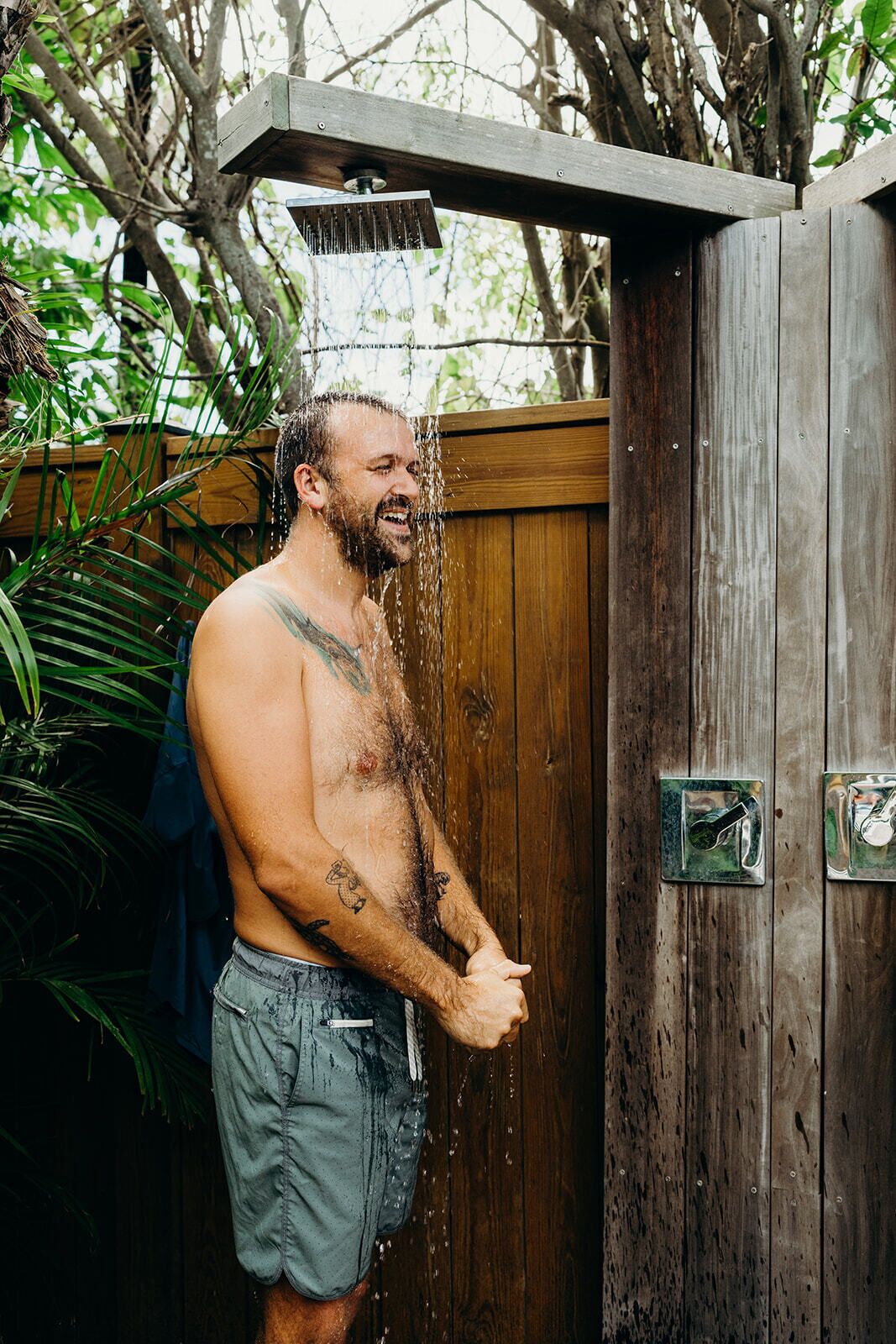 A man standing under a showerhead with water running, in front of a wooden structure with a metal handle.