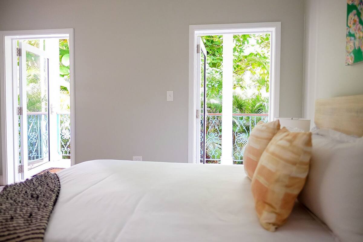 A bedroom with a bed, white walls, and a view of greenery through the window.