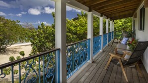 Una terraza de madera con barandillas azules, una zona de estar con una mesa y sillas, y vista a la playa y árboles.