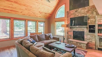 A cozy living room with a stone fireplace, a sectional sofa, and large windows offering a view of the outdoors.