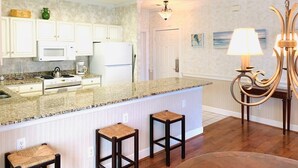 A kitchen with white cabinets, a granite countertop, a stove, and a refrigerator. There are two stools and a chandelier. - Magnolia North beautiful vacation condo (Myrtle Beach)