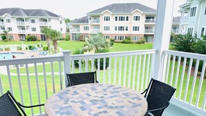 A covered outdoor area with a round table and four chairs, overlooking a pool and a building with multiple balconies. - Magnolia North beautiful vacation condo (Myrtle Beach)