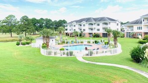 A residential building with a swimming pool surrounded by a white fence, lush green lawns, and trees. - Magnolia North beautiful vacation condo (Myrtle Beach)