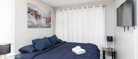 A bedroom with a dark blue bed, white curtains, a ceiling fan, a painting on the wall, and a TV mounted on the wall.