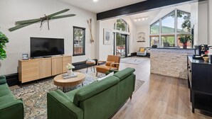 A spacious living room with a large TV, green sofas, and a dining area with a stone wall and large windows. - Lift House Lodge, Pet Friendly Studio Condo (Vail)