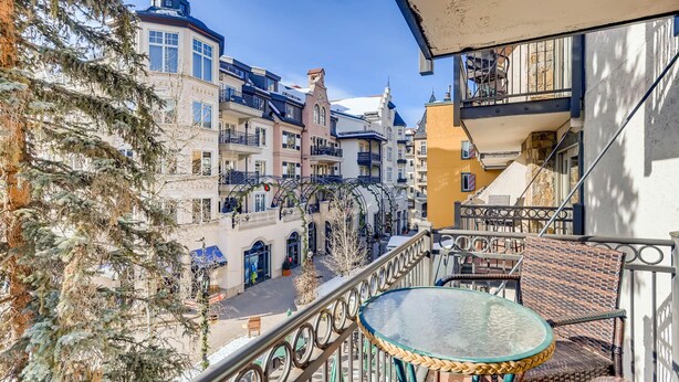 A balcony with wicker furniture, a glass table, and a view of a European-style street with buildings and a tree. - Lift House Lodge, Downtown Lionshead Remodeled Studio Condo (Vail)