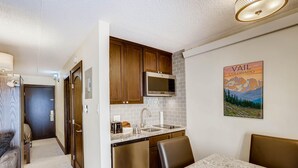 A compact kitchen with wooden cabinets, a stainless steel refrigerator, and a microwave. There is a dining area with a marble table and chairs. - Lift House Lodge, Downtown Lionshead Remodeled Studio Condo (Vail)