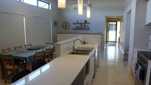 A modern kitchen with a central island, a dining area with wooden chairs, and a hallway leading to another room. - Anglers Rest at Sunderland Bay (Sunderland Bay)