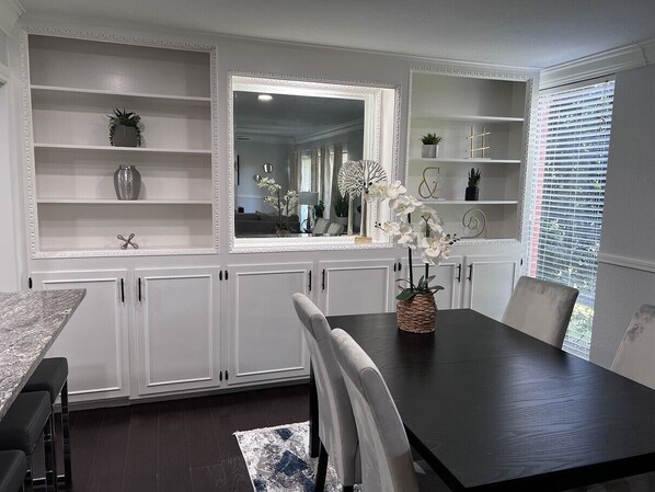 A dining area with a dark wooden table, white upholstered chairs, and a white cabinet with shelves. - GP36B | G. Prairie Condo | 3BD | Pool | Workspace (Grand Prairie)