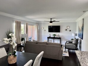 A living room with a sofa, dining table, chairs, a TV, and a ceiling fan. - GP36B | G. Prairie Condo | 3BD | Pool | Workspace (Grand Prairie)