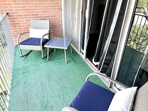 A balcony with a green artificial turf floor, a wicker chair with a cushion, a small table, and a sliding glass door. - GP36B | G. Prairie Condo | 3BD | Pool | Workspace (Grand Prairie)
