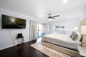A bedroom with a bed, a ceiling fan, a TV mounted on the wall, and a view of the outside.