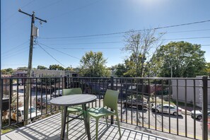 Un balcon avec une table et des chaises, donnant sur une rue bordée de voitures garées et d’immeubles.