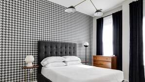 Un dormitorio con cabecera capitonada, cama con ropa de cama blanca, mesita de noche con lámpara, cómoda y papel tapiz estampado.